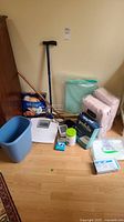 Photo showing assembled health aids including adult diapers, walking canes, scale, blood pressure monitor, trash bin, and other supplies on wooden floor near wall.