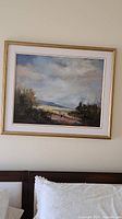 Framed oil painting of a landscape on canvas hung above a bed showing the full piece.