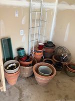 View of assorted clay, plastic, and metal planters stacked on the floor next to a white metal shelving unit in a garage.