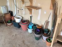 Wide view of garden planters and pots stacked against a wall in a garage, showing a variety of sizes, colors, and materials.