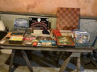 Wide shot of assorted board games and puzzles laid on a wooden bench including a checkerboard, Monopoly Canadian Edition, Ouija board, and various boxed games.