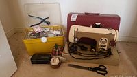 Full view of Lomain sewing machine beside red carrying case, yellow organizer box with sewing accessories, fabric scissors, pin cushions, and a leather hole punch tool.