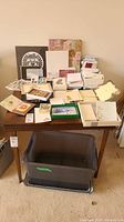 Full view of assorted stationery and card items on a wooden desk, showing multiple stacks of cards, envelopes, and decorative papers with a large plastic bin underneath