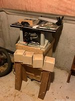 Full view of Delta table saw with metal stand, positioned on larger wooden base with wood boxes in front and other items around.