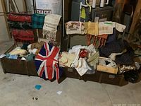 Two open vintage trunks on a basement floor, one filled with yarn skeins and spools, the other with various folded and draped vintage textiles including table runners and doilies.