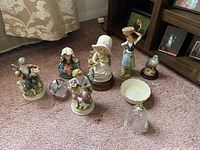 View of seven ceramic figurines, two decorative vases, and small bowl arranged on carpet in front of bookshelf and curtain.