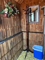 Wooden coat stand with twisted central post and four-legged base, placed in corner with wood-paneled walls and small trash bin.