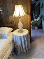 Crystal base table lamp with beige scalloped shade on round end table covered with lace and fabric cloth, set placed next to wooden paneled wall.