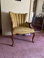 Front view of a large gold fabric upholstered armchair with scalloped backrest and curved wooden legs and arms.