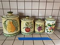 Four ceramic jars with fruit hand-painted designs lined up against a tiled wall