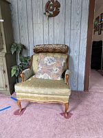 Front view of vintage green floral armchair with wooden arms and legs, featuring a paisley-patterned cushion and leopard-print neck pillow.
