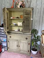 Front view of vintage wooden cabinet with upper shutter doors open, showing two interior shelves, single drawer below, and lower cupboard with doors closed. Distressed green paint with visible wear.