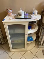 White wooden bathroom cabinet with a frosted glass door showing internal shelves, side curved shelving with three levels, and items placed on top for size and scale reference.