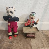 Front view of two vintage tin toys: a plush panda figure with red overalls and a Cragstan Carpenter tin toy figure with a workbench base.