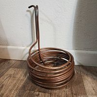 Photo showing full coiled copper wire coil with bent ends, standing against wall on wooden floor.