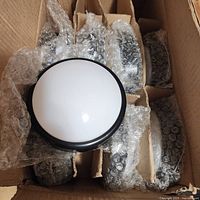 Top view of eight push lights in a cardboard box, each individually wrapped in bubble wrap. One light is unwrapped on top showing white dome and black base.