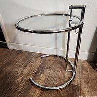 Full view of the adjustable side table showing round glass top and C-shaped chrome tubular base.