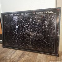 Full front view of a metallic Paris map wall decor on a floor leaning against a white wall showing detailed map in silver and black distressed style.