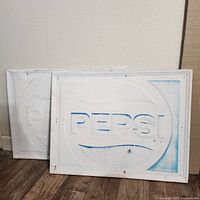 Two large metal Pepsi signs leaning against a wall on a wooden floor, showing embossed Pepsi logo and signs of wear.
