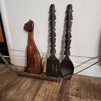 Wooden wall decor items: a carved cat figure with green eyes on left; two large carved utensils (fork and spoon) on the right, dark brown wood and hand-crafted details; all resting on wooden base with bow-shaped decoration.