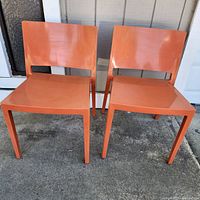 Front view of two orange plastic stacking chairs side by side on concrete ground, showing square seat and modern minimalist design.