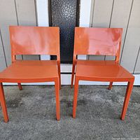 Two orange Kartell Lizz stacking chairs placed on concrete ground in front of a building exterior.