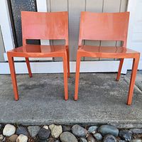 Front view of two orange molded plastic MCM Kartell Lizz stacking chairs placed side by side on cement surface, showing chair backs and seats.