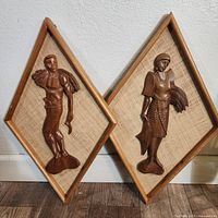 Front view of two mid-century diamond-shaped wooden wall art pieces with carved teak figures mounted on textured backing, showing detailed carving and wooden frame borders.