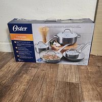 Front and side views of unopened Oster cookware set box showing product image, details in English and Spanish, and branding.