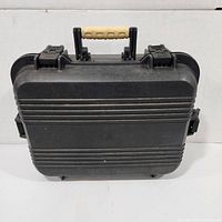 Plano hard plastic carrying case shown from front with beige grip handle and two latches, black exterior with horizontal ridges