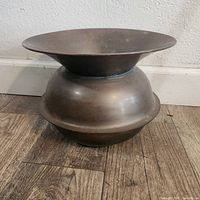 Side view of vintage brass spittoon showing bulbous body and flared rim on wooden floor against white wall.