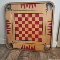 Photo of wooden square Carrom board side with red and cream checkerboard pattern and red triangular designs. Board shows wear.