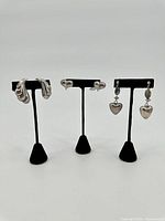 Three pairs of sterling silver earrings on display stands: abstract wavy hoops, heart studs, and heart dangle earrings with beads.