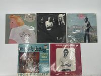 Five vinyl records in their sleeves arranged on a flat surface showing album cover art with a variety of styles and artists.