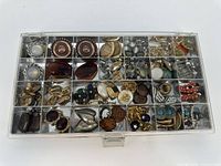 Full view of all compartments in a clear plastic jewelry organizer showing an assortment of costume jewelry pieces including earrings, brooches, and rings in mixed metals and colors