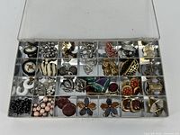 Top down view of clear plastic box showing multiple small compartments each containing various pieces of costume jewelry including earrings, clips, and brooches in varied colors and materials.