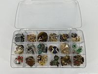 Clear plastic compartment box filled with assorted costume jewelry including earrings, rings, pins, and pendants in varied colors and metals.