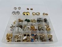 Overview photo showing multiple compartment organizer tray filled with an assortment of costume jewelry primarily earrings and pins in gold and silver tones with various designs.