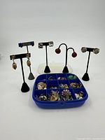 Display of multiple pairs of earrings on black stands and a blue storage box containing many pairs of earrings and costume jewelry pieces separated into compartments.