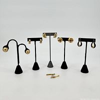 Front view of five pairs of gold-colored Napier earrings displayed on black stands; shows a variety of styles including studs, hoops, and clip-ons.