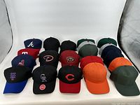 Photo showing 20 baseball caps arranged in five rows of four caps each, including a mix of MLB team logos and plain caps in various colors.