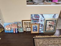 Wide view of various framed artwork and prints lined up on floor, including large canvas print of coastal cityscape, Mona Lisa print, Egyptian-themed artwork, and smaller pieces.