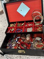 Open jewelry box showing a variety of earrings and bangles in red fabric-lined compartments.