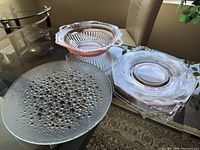 Three glassware items on glass table by window: two pink depression glass pieces and one bubbly clear plate with colorful bubbles