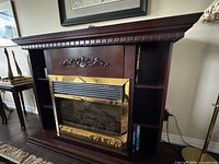 Front and angled view of the electric fireplace showing dark wood casing, gold-tone metal trim, and side shelves with books. Visible repair on top left corner and marks near base.