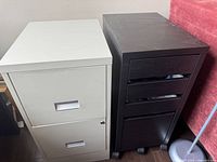 Photo showing off-white metal file cabinet with two drawers alongside dark wood finish multi drawer cabinet on wheels in room interior.