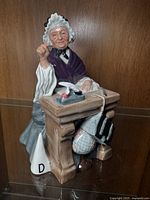 Frontal-side view of the School Marm figurine showing the seated elderly woman with a white bonnet and purple shawl, with the desk and dunce cap visible.