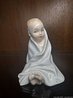 Front view of Royal Doulton porcelain figurine, 'This Little Pig', showing seated child wrapped in blanket with visible feet and smiling face.