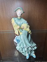 Side view showing detailed ruffled green dress, seated woman holding a yellow fur stole, and green hat with blue flowers on a brown chair.