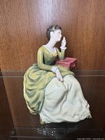 Front side view of Royal Doulton Carolyn figurine seated on glass shelf showing detailed face, gown with green and cream colors, and pink side table
